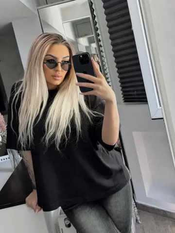 Blonde woman in a black t-shirt and grey jeans taking a stylish mirror selfie in a modern room.