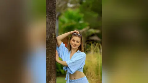 Smiling woman in a light blue linen crop top leaning against a tree in a lush green garden.