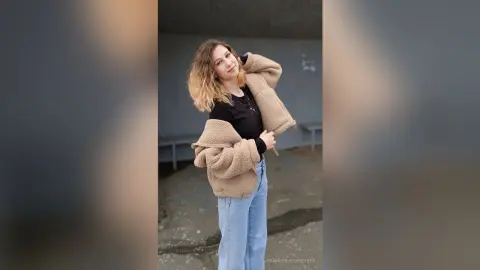 Smiling woman in a cozy beige fleece jacket and light blue jeans standing in an urban outdoor setting.