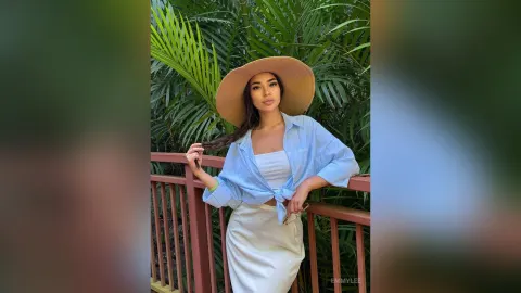 EmmyLee — EmmyLee wears a wide-brimmed straw hat and a light blue tied shirt while leaning against a wooden railing in a tropical garden.