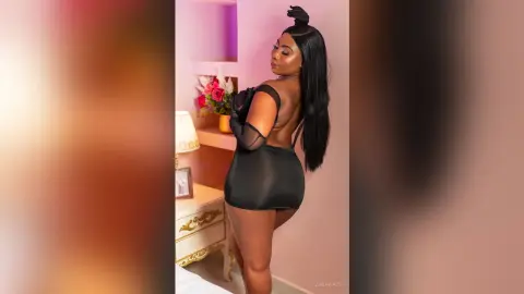 Elegant woman in a black sheer mini dress posing in a soft pink bedroom setting.