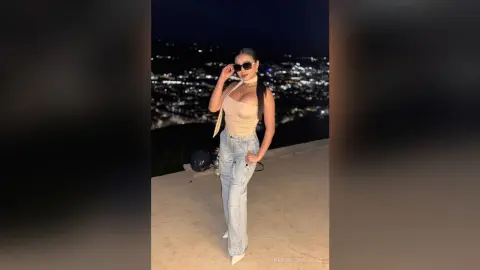 Stylish woman in beige halter top and light wash jeans posing against a glowing city night skyline.
