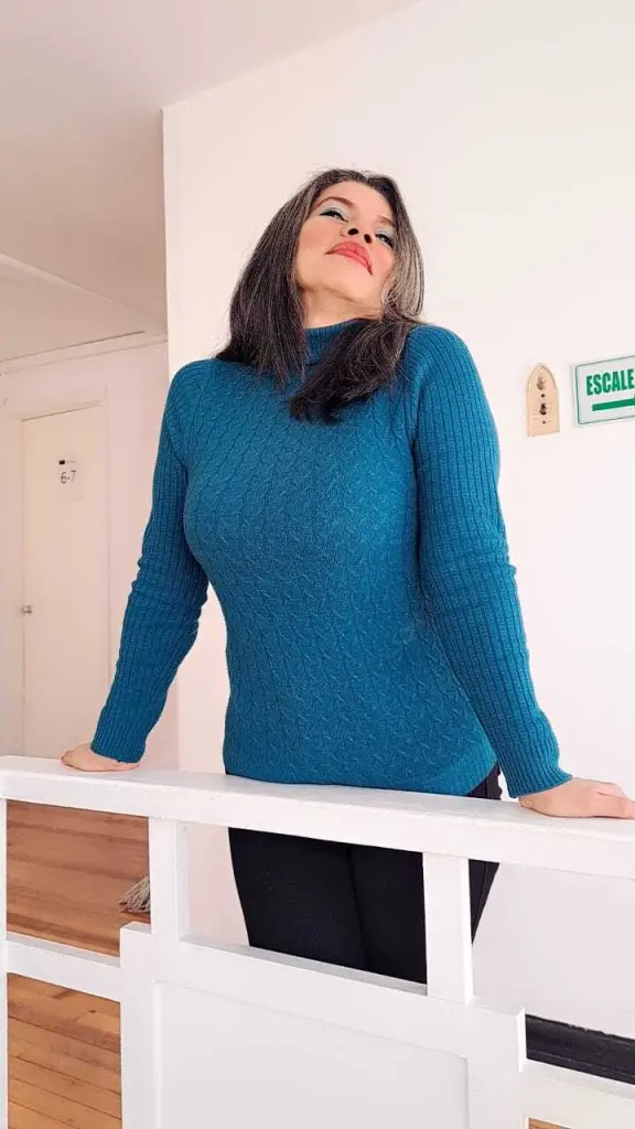 VikyAdams72 with long grey hair leans her chin on her hand while wearing a blue long-sleeved ribbed top.
