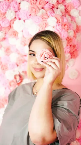 Blonde woman in a grey t-shirt holding a pink rose in front of her eye against a floral rose wall background.