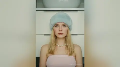 Blonde woman in a light blue faux fur hat and pink strapless top posing in a bright, minimalist room.