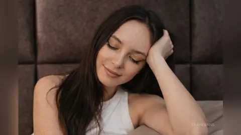 LeahEvans rests her head on her hand with her eyes closed, wearing a white tank top against a dark headboard.