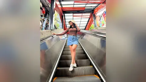 Stylish woman in a brown top and denim skirt posing on a modern escalator under a bright red canopy.