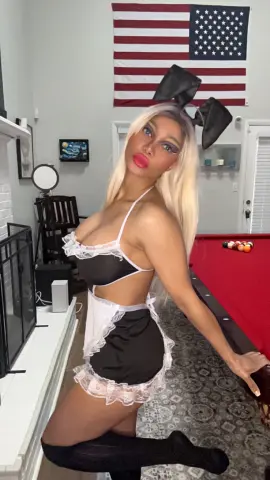 Blonde woman in a black and white maid cosplay outfit posing in a living room with an American flag.