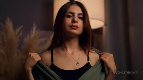 Young woman with nose piercing wearing a black tank top and green cardigan in a moody indoor setting.