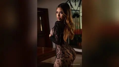 Glamorous woman in a black sheer patterned dress posing in a moody, dimly lit indoor setting.