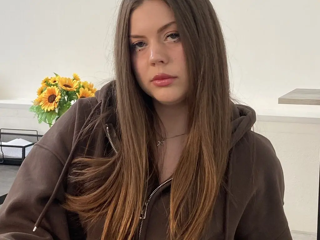 HildredBrage wearing a brown zip-up hoodie with her long brown hair down, posing against a white wall with sunflowers in the background.