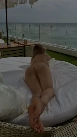 Blonde woman relaxing on a white outdoor daybed overlooking the ocean at a tropical seaside resort.
