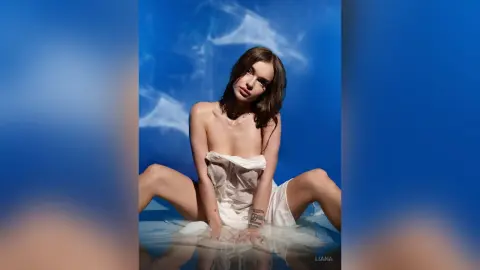 Elegant woman in a white draped dress sitting against a dreamy blue cloud background.