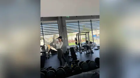 NicoleBella stands in a gym wearing a white crop top and black leggings, taking a mirror selfie next to a weight bench and dumbbells.