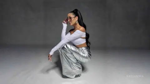 Stylish woman in white crop top and silver metallic trousers posing confidently against a grey studio background.