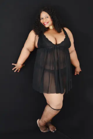 Curvy woman in a black sheer lace chemise and fishnet stockings posing against a dark studio background.