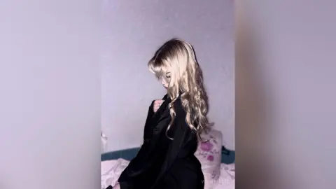 Blonde woman in a black outfit posing with long wavy hair in a soft, moody indoor setting.