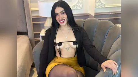 Smiling woman in a black blazer and gold skirt sitting on a grey velvet shell chair in a bright room.