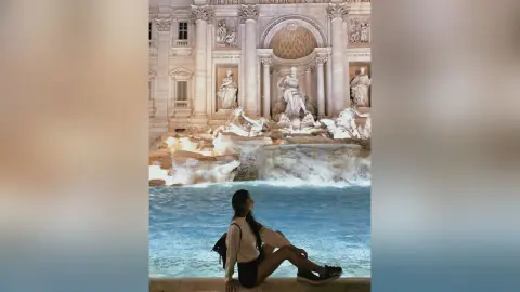 LucyMartin sits on the edge of a stone fountain in a white top and black shorts, looking back over her shoulder.