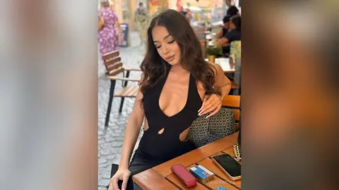 Brunette woman in a black halter bodysuit sitting at an outdoor cafe table on a sunny day.