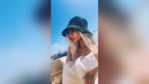 Blonde woman in a black bucket hat and white top posing under a bright blue sky on a sunny balcony.