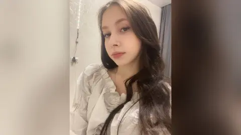Young woman with long wavy hair wearing a white ruffled blouse in a soft, minimalist indoor setting.