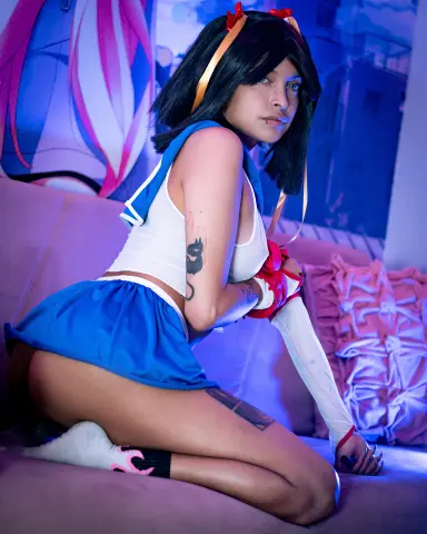 SakuraTao poses on a pink couch wearing a white tank top and blue skirt, showing off her black hair and arm tattoos.