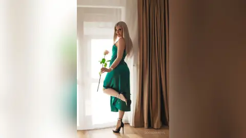 Elegant blonde woman in a green satin dress holding a rose by a bright window in a dreamy indoor setting.