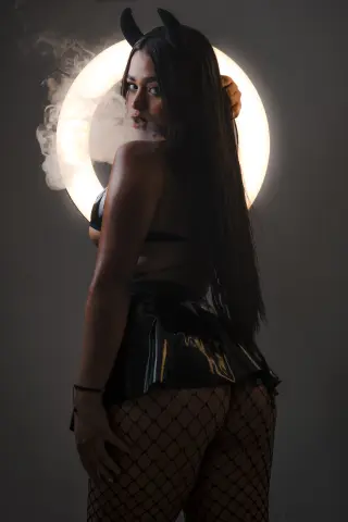 Mysterious woman in black devil cosplay with horns, leather skirt, and fishnets against a glowing halo light.