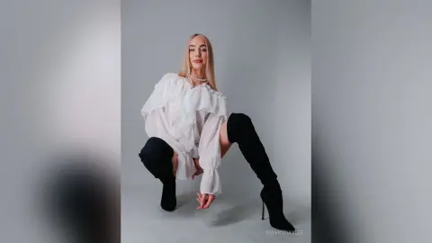 MarryDavies poses in a white ruffled blouse and black thigh-high boots against a grey background.