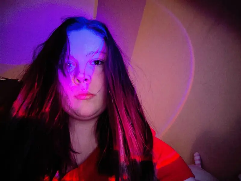 chillnsmoke poses with her long dark hair falling over her face and shoulders, wearing a black top against a bright background.