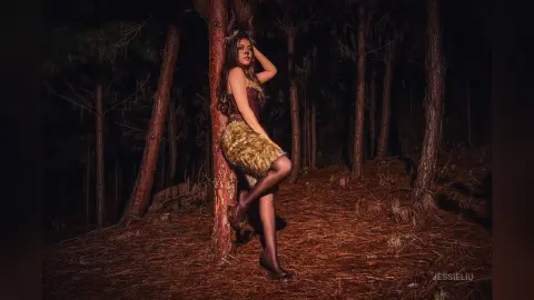 JessieLiu leans against a tree trunk in a dark forest wearing a gold fringed skirt and black hosiery.