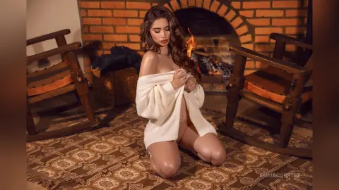 Woman in a white knit sweater sitting on a patterned rug by a cozy brick fireplace.