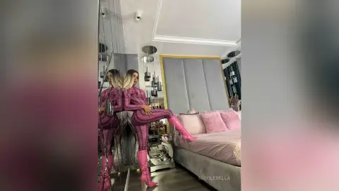 NicoleBella poses in a pink patterned bodysuit and matching pink boots while standing next to a bed in a modern room.