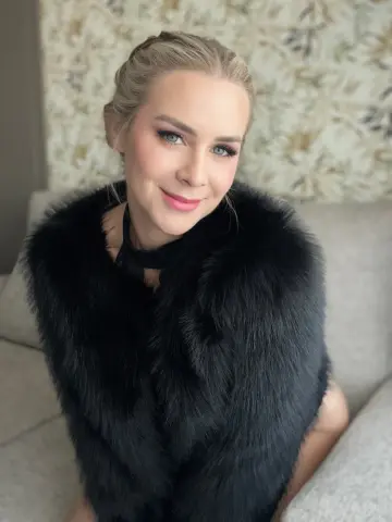 Elegant blonde woman in a luxurious black faux fur coat sitting on a sofa in a bright room.
