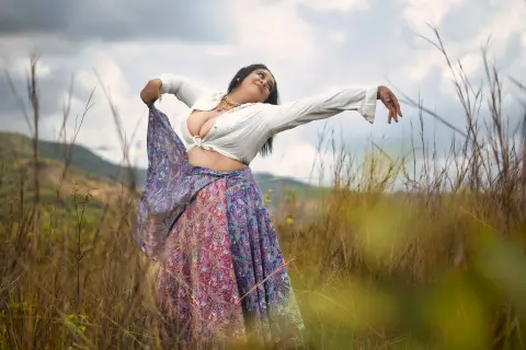 Woman in white blouse and colorful patterned skirt dancing gracefully in a grassy field under a cloudy sky.