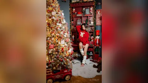 Young woman in a red sweater and white fuzzy socks posing by a decorated Christmas tree in a festive room.