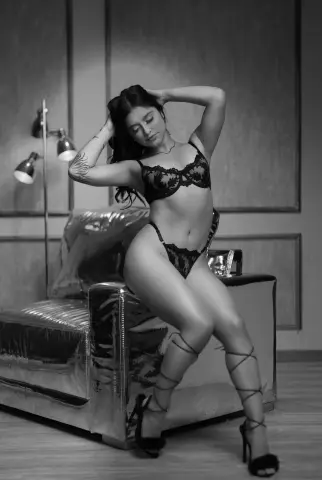 IndiraJhons poses sitting on a silver metallic chair wearing black lace lingerie and strappy high heels.