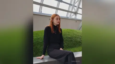 Redheaded woman in a black turtleneck and trousers sitting on a bench in a modern glass building.