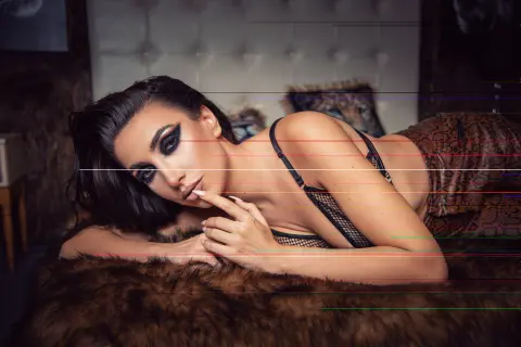 Glamorous woman in black lace lingerie lounging on a faux fur rug in a moody, dimly lit bedroom setting.