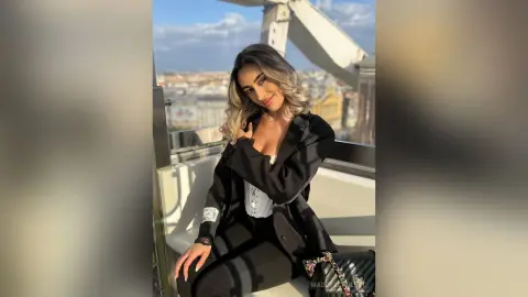 Smiling woman in a chic black blazer and white top posing on a sunny urban balcony overlooking a city skyline.