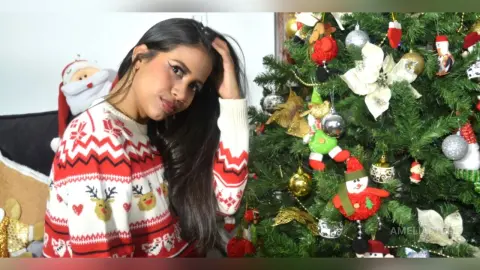 AmeliaStell wears a red and white patterned Christmas sweater while posing next to a decorated Christmas tree.