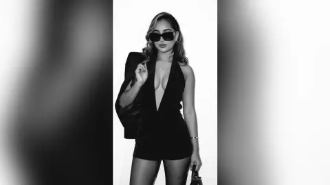 Stylish woman in a black plunging jumpsuit and sunglasses posing against a white background.
