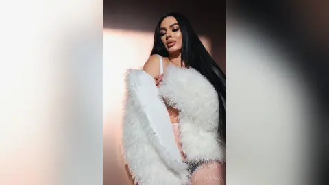 Glamorous woman in a white faux fur coat and lace bodysuit posing against a soft pink studio background.