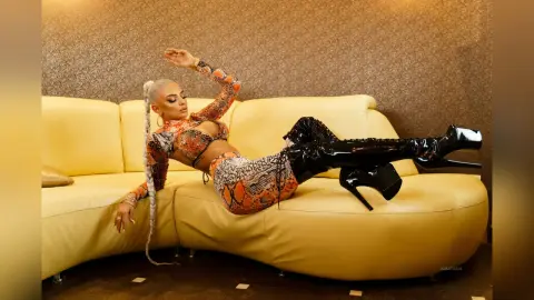 Anastasia reclines on a yellow leather sofa wearing an orange patterned bra and matching bottoms with black thigh-high boots.