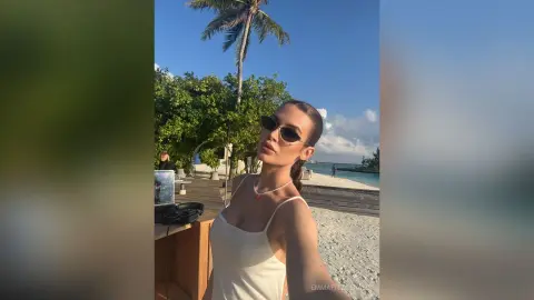 Stylish woman in a white tank top and sunglasses relaxing on a sunny tropical beach near palm trees.