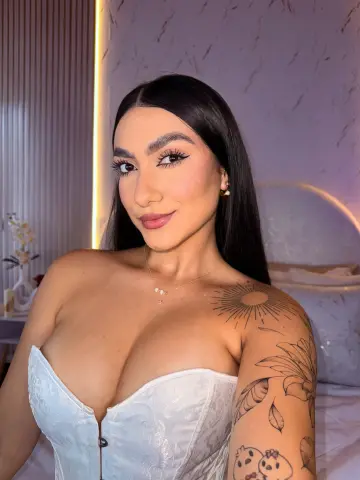 Young woman with long dark hair in a white corset posing in a cozy bedroom setting.