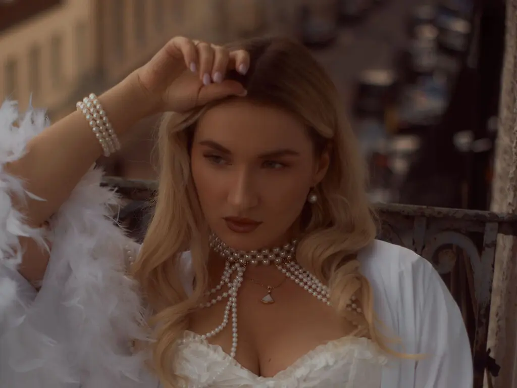Elegant blonde woman in a white feathered outfit and pearl necklace posing on a balcony with a dreamy mood.