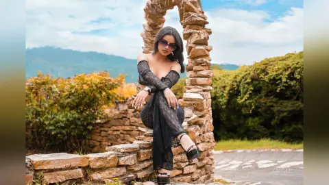 Stylish woman in a black lace dress and sunglasses posing by a rustic stone archway in a scenic mountain landscape.