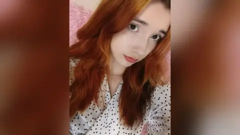 Redheaded woman in a white polka dot blouse smiling softly in a cozy pink setting.